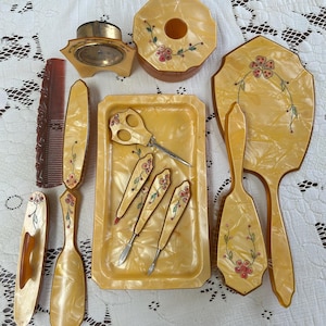 May include: A vintage vanity set in shades of yellow and cream, featuring a hand mirror, brush, comb, tray, and various manicure tools. Each piece is decorated with floral accents in pink and green.