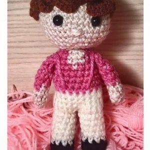 May include: A handmade crochet doll with a brown hair, black eyes, and a pink sweater. The doll is wearing white pants and black shoes. It is sitting on a bed of pink yarn.