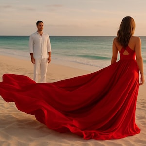 Peut inclure: Une femme porte une longue robe rouge sur une plage de sable, face à la mer. Un homme en vêtements blancs se tient à proximité. La robe a un dos croisé et une longue traîne. Le ciel est orange et l'eau turquoise. Scène au coucher du soleil.