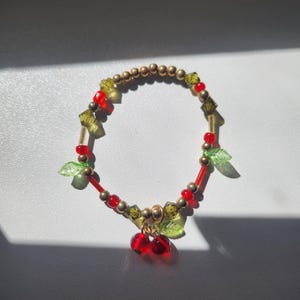 Könnte beinhalten: Ein rotes und grünes Perlenarmband mit einem Kirsch-Anhänger. Das Armband besteht aus kleinen goldenen Perlen, roten und grünen Glasperlen und zwei grünen Blatt-Anhängern. Der Kirsch-Anhänger besteht aus roten Glasperlen und hat einen goldenen Stiel.