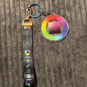 May include: A black fabric keychain strap featuring white graphic icons and a rainbow-coloured circular acrylic charm. The strap includes a metal clasp and a split ring, with a design displaying various symbols and a colour wheel gradient.