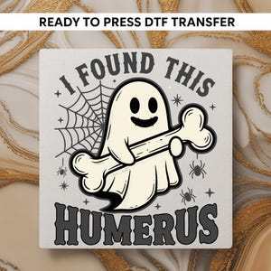 May include: A square, light gray DTF transfer with a Halloween theme. The design features a cartoon ghost holding a bone, with the text "I FOUND THIS HUMERUS". A spider web, spiders, and stars complete the design.