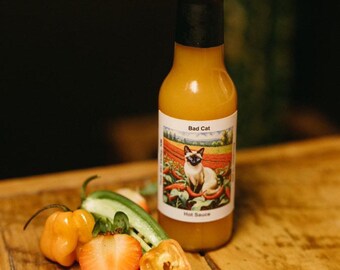 Könnte beinhalten: Eine Glasflasche mit gelber scharfer Sauce mit schwarzem Verschluss und einem Etikett mit der Aufschrift "Bad Cat Hot Sauce". Das Etikett zeigt eine Karikatur einer siamesischen Katze, die auf einer roten Pfefferpflanze sitzt. Die Flasche steht auf einem Holztisch mit einer gelben Habanero-Pfeffer, einer grünen Gurke und einer geschnittenen Erdbeere.