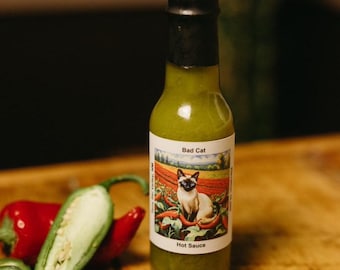 Op de afbeelding: Een glazen flesje met groene hete saus met een zwarte dop en een etiket met de tekst "Bad Cat Hot Sauce". Het etiket toont een cartoon van een Siamese kat die in een veld met rode pepers zit.