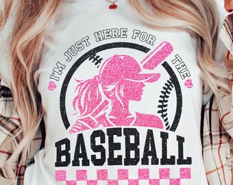 Może przedstawiać: Biała koszulka z różowo-czarnym nadrukiem graficznym. Grafika przedstawia kobietę grającą w baseball z kijem i tekstem "I'm just here for the baseball".