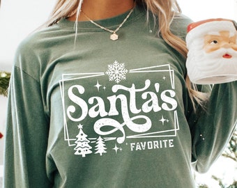 May include: Sage green long-sleeve shirt with the text "Santa's Favorite" in a white, festive design. The shirt features a snowflake, Christmas trees, and stars. A Santa mug is held in the hand. The shirt is part of a Christmas theme.