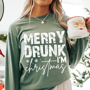 Puede incluir: Camiseta de manga larga verde salvia con el texto "MERRY DRUNK I'M christmas" en blanco, con fuente desgastada. La camiseta presenta copos de nieve y un "christmas" en cursiva. Se sostiene una taza de Papá Noel.