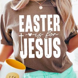 Puede incluir: Una camiseta marrón con el texto "EASTER is for JESUS" en blanco. La camiseta se combina con pantalones cortos turquesa y una taza blanca con interior amarillo y una carita sonriente.
