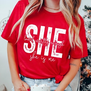 May include: Red t-shirt with a white graphic that says "She loved worthy enough she is me". The graphic has a distressed look. The shirt is being worn by a person holding a mug that says "Sweater Weather".