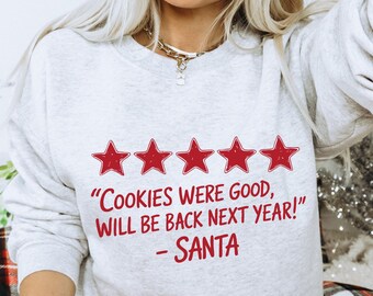 以下が含まれることがあります： 「COOKIES WERE GOOD, WILL BE BACK NEXT YEAR! - SANTA」の赤い文字がプリントされたライトグレーのスウェットシャツ。テキストの上には5つの赤い星があります。ストロー付きの赤いタンブラーが手に持たれています。