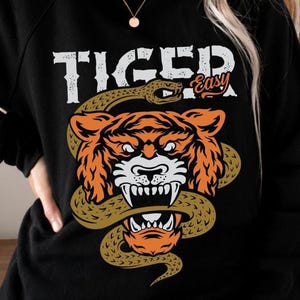 Könnte beinhalten: Schwarzes Sweatshirt mit einem Aufdruck eines Tigerkopfes, der von einer Schlange umwickelt wird. Der Text "Tiger Easy" ist über dem Aufdruck gedruckt.