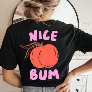 May include: Black t-shirt with a peach graphic and the words "NICE BUM" in pink. The peach is orange with green leaves. The shirt is made of a soft material.