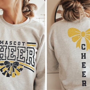 Puede incluir: Una sudadera gris con las palabras "MASCOT CHEER" en negro con un lazo de purpurina dorada en la parte delantera. La parte posterior de la sudadera tiene un lazo de purpurina dorada y la palabra "CHEER" verticalmente en negro.