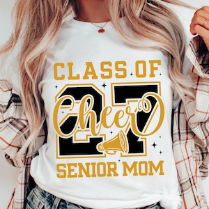 Puede incluir: Camiseta blanca con un gráfico dorado y negro que dice "CLASS OF 27 CHEER SENIOR MOM". El diseño incluye un megáfono y estrellas. La camiseta es de cuello redondo.
