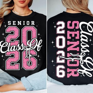 Könnte beinhalten: Schwarzes Sweatshirt mit dem Text "SENIOR 2026 Class Of" in weißer und rosa Glitzer-Schrift auf der Vorderseite. Die Rückseite des Sweatshirts hat den gleichen Text in vertikaler Anordnung. Das Design beinhaltet eine Schleife.