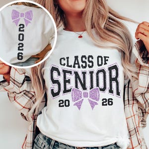 Op de afbeelding: Wit T-shirt met een glitterende paarse strik en de tekst "CLASS OF SENIOR 2026". De achterkant van het shirt heeft een bijpassende strik en het nummer "2026" in zwart.