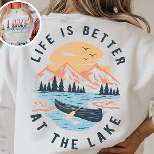 May include: White sweatshirt with a scenic lake graphic. The design features a sunset, mountains, trees, a boat, and the text "LIFE IS BETTER AT THE LAKE". The front of the shirt has a smaller graphic with the text "LAKE Junkie".