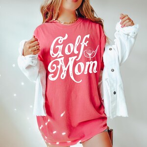 Pu&ograve; includere: Una maglietta rosa con il testo "Golf Mom" in bianco con una grafica di una mazza da golf e una palla.