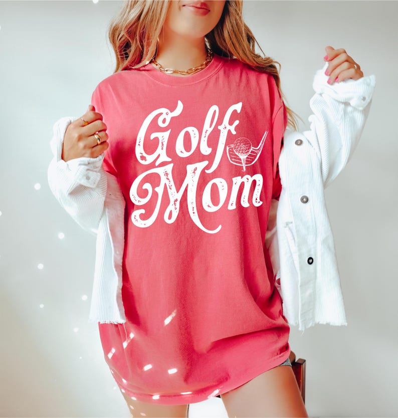 Pu&ograve; includere: Una maglietta rosa con il testo "Golf Mom" in bianco con una grafica di una mazza da golf e una palla.