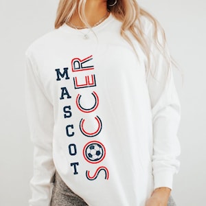 Op de afbeelding: Wit shirt met lange mouwen met de woorden "MASCOT SOCCER" verticaal gedrukt in marineblauw en rood. Het woord "SOCCER" bevat een voetbalafbeelding. Een casual kledingstuk.