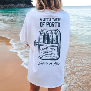 May include: White t-shirt with a graphic of a sardine can, featuring the text "A LITTLE TASTE OF PORTO" and "SARDINES WILD CAUGHT IN OLIVE OIL". The shirt also includes the phrase "Delicia do Mar".