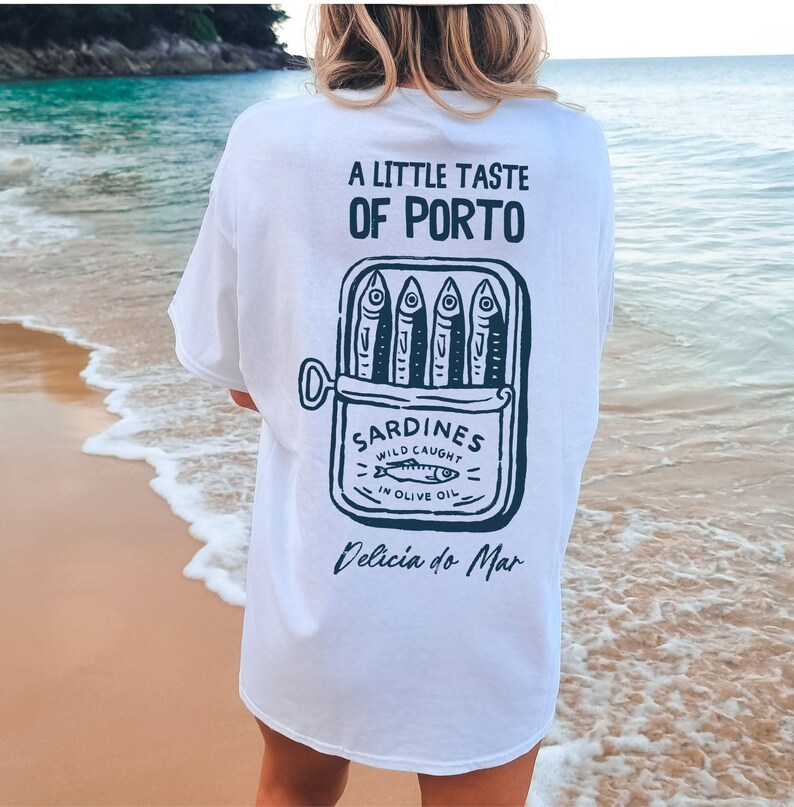 May include: White t-shirt with a graphic of a sardine can, featuring the text "A LITTLE TASTE OF PORTO" and "SARDINES WILD CAUGHT IN OLIVE OIL". The shirt also includes the phrase "Delicia do Mar".