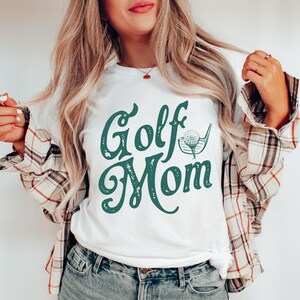 Pu&ograve; includere: Maglietta bianca con una grafica verde in stile vintage che dice "Golf Mom" con un'illustrazione di una palla da golf e una mazza.