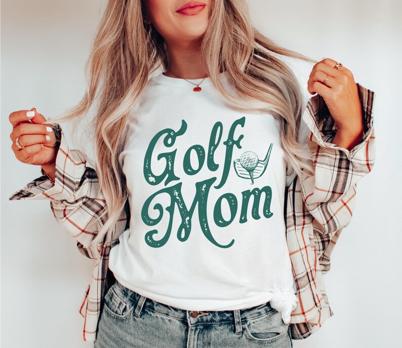 Pu&ograve; includere: Maglietta bianca con una grafica verde in stile vintage che dice "Golf Mom" con un'illustrazione di una palla da golf e una mazza.