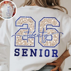 Op de afbeelding: Wit sweatshirt met gouden glitter 2026 en het woord "twenty" in cursief. Het woord "SENIOR" staat in blauw onder de cijfers.