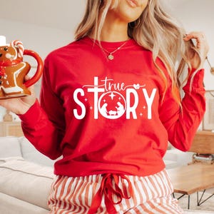 May include: A red long-sleeve shirt with the text "True Story" in white, featuring a cross and a nativity scene graphic. A gingerbread man mug with a white top and red scarf is held in the hand.