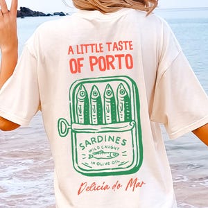 May include: Cream-colored t-shirt with a graphic of a sardine can, featuring the text "A Little Taste of Porto" in red and "Sardines Wild Caught in Olive Oil" in green. The phrase "Delicia do Mar" is written in red.