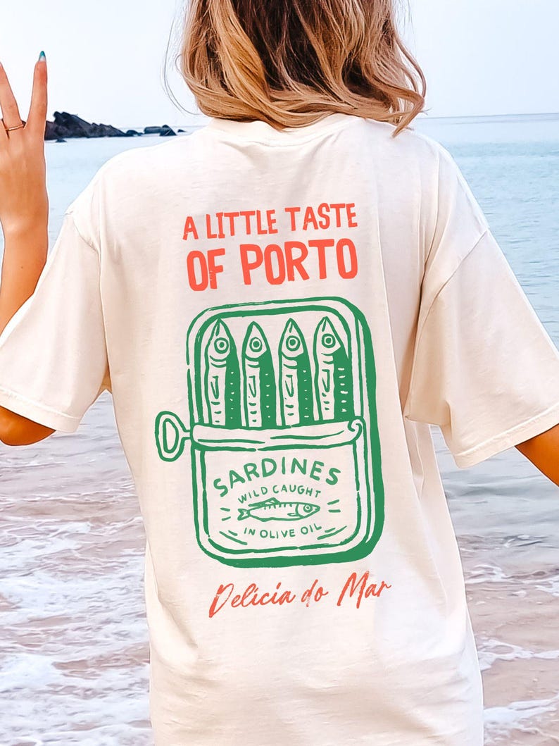 May include: Cream-colored t-shirt with a graphic of a sardine can, featuring the text "A Little Taste of Porto" in red and "Sardines Wild Caught in Olive Oil" in green. The phrase "Delicia do Mar" is written in red.