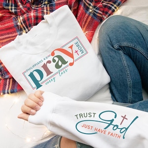 Peut inclure: Deux sweats blancs avec des messages religieux. Le premier sweat-shirt a le texte "Prier plus, s'inquiéter moins" avec le verset biblique "Philippiens 4:6" en dessous. Le deuxième sweat-shirt a le texte "Faire confiance à Dieu, il suffit d'avoir la foi" avec une croix.
