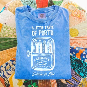 May include: A blue t-shirt with the text "A Little Taste of Porto" and a graphic of sardines in a can. The shirt also has the text "Sardines Wild Caught in Olive Oil" and "Delicia do Mar".