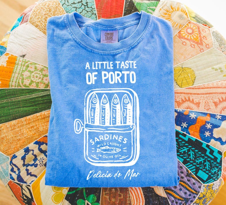 May include: A blue t-shirt with the text "A Little Taste of Porto" and a graphic of sardines in a can. The shirt also has the text "Sardines Wild Caught in Olive Oil" and "Delicia do Mar".