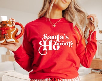 May include: A red long-sleeve shirt with the text "Santa's Favorite Ho" in white script. A gingerbread man mug with a white marshmallow top and candy cane handle is held in the hand. The mug is brown with a red scarf.