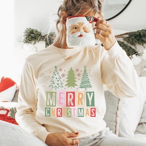 Puede incluir: Camiseta de manga larga color crema con un diseño de árbol de Navidad y el texto "MERRY CHRISTMAS" en letras de bloque coloridas. Una taza de Papá Noel se sostiene frente a la cara. Ropa festiva navideña.