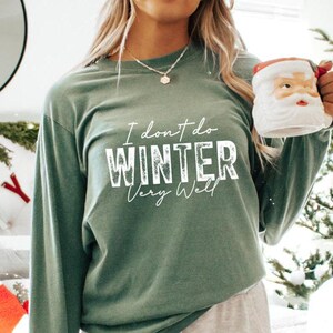 Puede incluir: Una camiseta verde de manga larga con el texto "I don't do winter very well" en letras blancas. La camiseta la lleva puesta una persona que sostiene una taza blanca con la cara de Pap&aacute; Noel.