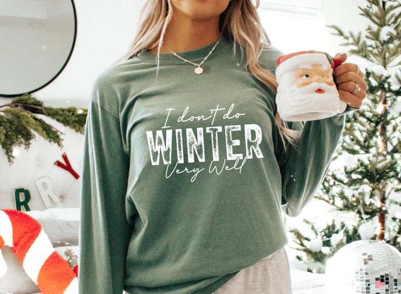 Puede incluir: Una camiseta verde de manga larga con el texto "I don't do winter very well" en letras blancas. La camiseta la lleva puesta una persona que sostiene una taza blanca con la cara de Pap&aacute; Noel.