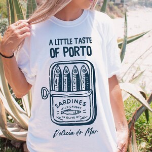 May include: White t-shirt with a graphic of a sardine can, featuring the text "A Little Taste of Porto" and "Sardines Wild Caught in Olive Oil." The shirt also includes the text "Delicia do Mar."