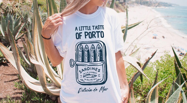 May include: White t-shirt with a graphic of a sardine can, featuring the text "A Little Taste of Porto" and "Sardines Wild Caught in Olive Oil." The shirt also includes the text "Delicia do Mar."