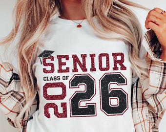 May include: White t-shirt featuring the text "SENIOR CLASS OF 26" in a glittery maroon font and black outline. A graduation cap graphic is above the word "SENIOR". The shirt is worn by a person with blonde hair and a plaid shirt.