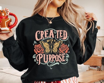 May include: Black sweatshirt with the text "CREATED WITH A PURPOSE" in pink and teal. A yellow butterfly and two pink roses are featured in the design. The sweatshirt is a casual, comfortable style.