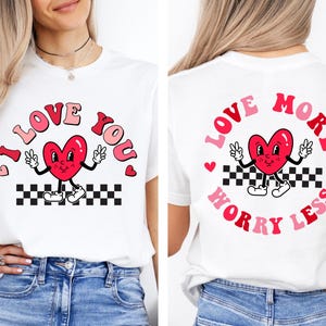 May include: White t-shirt featuring a cartoon heart with arms and legs, peace signs, and the words "I Love You" in pink and red. The design includes a black and white checkered pattern at the bottom. The back of the shirt says "Love More Worry Less".