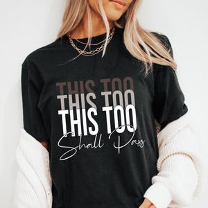 May include: Black t-shirt with the text "This Too Shall Pass" in white lettering with a gradient effect in shades of brown.