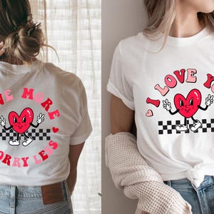 May include: White t-shirts with a retro design featuring a red heart character with peace signs. One shirt says "Love More Worry Less" in a circle, the other says "I Love You". Both have a black and white checkered pattern.