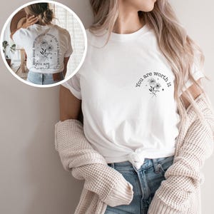 May include: White t-shirt with the text "You are worth it" and a floral design on the front. The back of the shirt has the text "Keep growing every day is a fresh start you are worth it" with a floral design.
