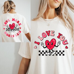May include: Cream-colored t-shirt featuring a retro design with a red heart character giving peace signs. The words "I LOVE YOU" are arched above the heart. A matching design is on the back, with the words "LOVE MORE WORRY LESS".