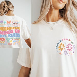 Puede incluir: Una camiseta blanca con un diseño gráfico colorido en la parte delantera y trasera. El diseño delantero presenta dos flores estilizadas con el texto "Normalizar las mentes de todo tipo". El diseño trasero presenta el texto "Normalizar las mentes de todo tipo" con una variedad de ilustraciones y textos coloridos, incluyendo "Eres suficiente", "No seas tan duro contigo mismo" y "Acabar con el estigma".
