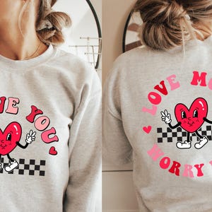 May include: A light gray sweatshirt with a retro design. The front features the text "I LOVE YOU" with a cartoon heart character. The back has "LOVE MORE WORRY LESS" with a similar heart character. The design includes pink, red, black, and white.